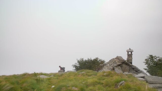 Lone hiker with backpack hiking on misty alpine mountain meadow near stone hut in foggy summer weather, outdoor adventure trekking in the european alps