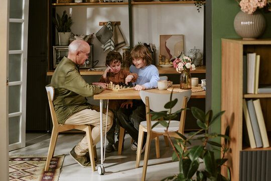 Senior Caucasian man playing chess with two kids at home, teaching strategy and bonding across generations. Useful for family lifestyle, parenting, retirement, education campaigns