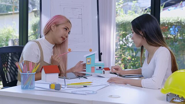 Creative female engineers brainstorming on a residential building project using a tablet and house model in a bright workspace.