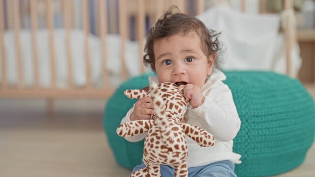 Toddler girl chews and holds a stuffed giraffe toy while sitting by a crib in a building; playful curiosity.