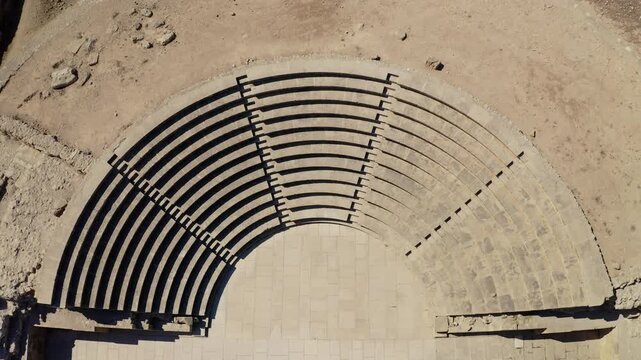 Archaeological site of a historic Roman Odeon in the ancient city of Paphos, Cyprus, featuring stone seating tiers and an arena, viewed from an aerial perspective on a sunny day. Drone flight zoom out