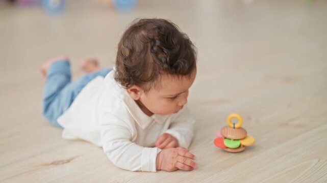 Hispanic baby holding colorful wooden teething toy with hands and mouthing it on a light wood floor in studio; curiosity bonding.