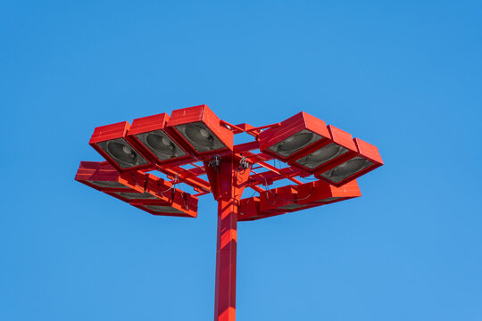 Red stadium floodlights fill the frame against a cloudless blue sky. The close angle shows metal housings, lenses, cables, and urban lighting detail.