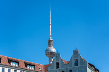 The Berlin TV Tower rises behind tiled rooftops and stepped gables. Its silver sphere and striped antenna stand out against a vivid blue sky. © Jakob