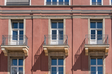 Obraz premium Pink residential facade shows balconies, windows and sharp sunlit shadows. Small shrubs appear at the base of the urban building.