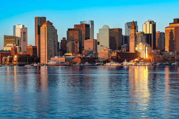 Obraz premium A wide view across the bay from East Boston captures the downtown Boston skyline at sunrise with the water reflecting warm golden light on the city.