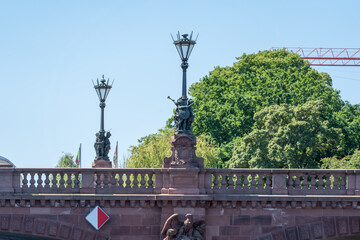 Obraz premium Decorative lanterns and sculpted figures top a stone bridge above the Spree in Berlin. Leafy deciduous trees and a crane frame the riverside landmark.