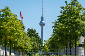 The Berlin TV Tower stands between leafy deciduous trees with a red flag nearby. The sunny city view combines modern landmark architecture with summer greenery. © Jakob