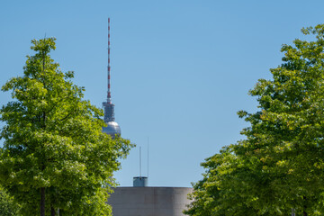 The Berlin TV Tower rises beyond green deciduous trees and modern architecture. Clear blue sky and summer foliage frame the famous city landmark. © Jakob