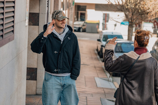 Young man in cap and jacket talking with a woman on an urban sidewalk, both gesturing expressively during a casual street conversation