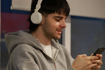 young man wearing white headphones and a gray hoodie smiles while using his phone in a bright,...