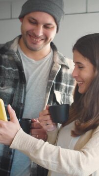 Video of a young Caucasian couple on a winter day in their home kitchen enjoying a cup of coffee while watching videos on their mobile phone. Concept of a winter day at home, young marriage, and famil