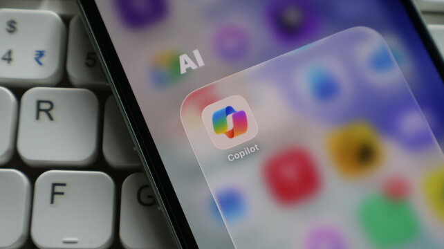 Closeup of a smartphone screen displaying the microsoft copilot app icon inside an ai folder, with a computer keyboard in the background