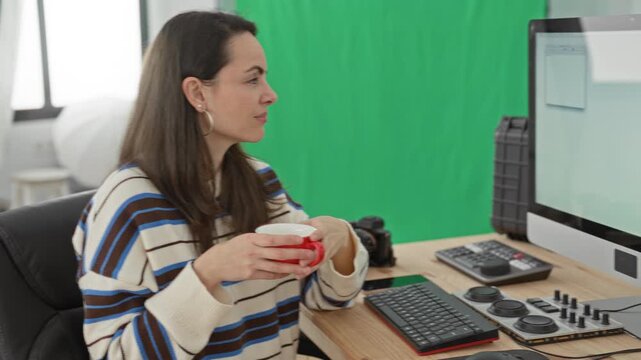 Woman holds red coffee cup with both hands and looks at computer monitor in a studio with green screen; calm concentration.
