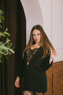 A young woman in a black blazer and lace bodysuit stands near an arch posing playfully, touching her hair and pouting at the camera