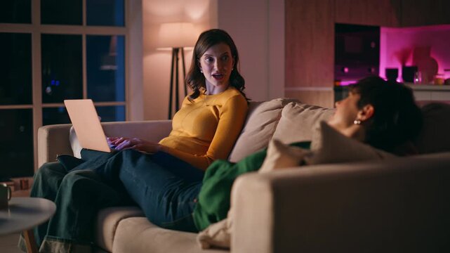Relaxed roommates enjoy domestic leisure in evening home. Woman browsing laptop