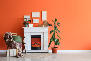 Fototapeta premium Fireplace with armchair, houseplants and photo frames near orange wall in living room