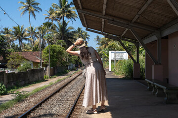 Obraz premium A scenic lifestyle view of a young Caucasian female tourist in a straw hat and long skirt standing on an empty concrete platform of a rural railway station surrounded by palm trees