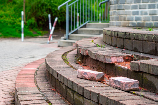 Damaged circular outdoor stairs made of paving bricks with visible defects and loose blocks, photographed in daylight from a low perspective with a blurred background showing metal railing and greener