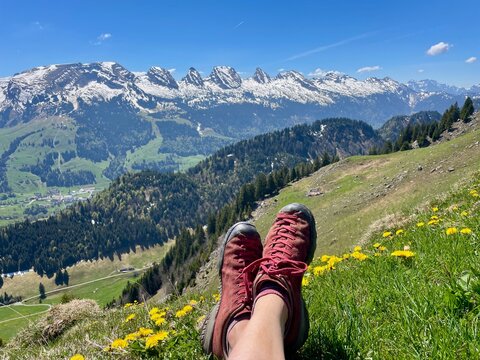 Feet wearing hiking boots resting in front of the panoramic view of the Churfirsten mountain range in the Swiss Alps, Toggenburg region, St. Gallen, Switzerland.