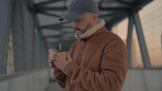 He is holding a paper coffee cup with both hands, taking a warm sip in the chilly urban air. The sharp depth of field perfectly separates the subject from the geometric metal structures blurring.