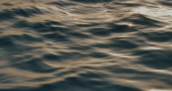 Close-up cinematic shot of calm ocean water with soft waves and warm golden sunset reflections. Natural abstract background with relaxing motion and tranquil atmosphere.
