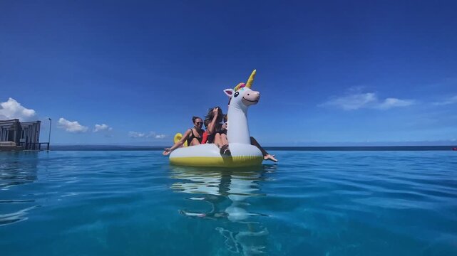 Friends afloat on unicorn float near overwater bungalow, panoramic turquoise ocean and cloudless sky, tranquil travel pause with gentle ripples and distant resort silhouette, Bali island