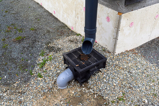 A black plastic drainage grate embedded in gravel collects rainwater from a dark downpipe; a grey outlet pipe extends underground beside a concrete foundation wall