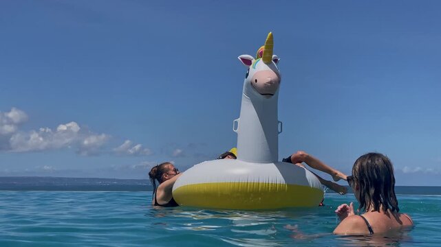 Two women and two boys playing on inflatable unicorn float in turquoise water under clear blue sky, friends laughing and splashing, lounging on giant horned float, sunny relaxed, Bali island