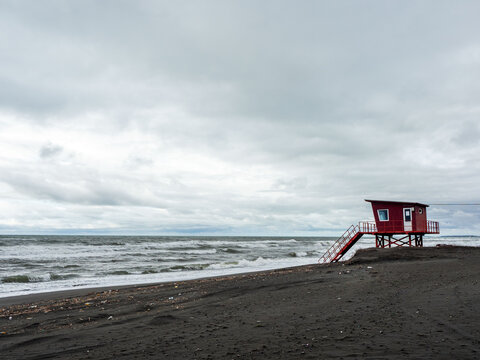 side view of a red wooden lifeguard station on a dark sand beach during a gloomy day. The stairs lead down to a stormy ocean with white waves under a dramatic overcast sky