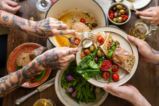 Tattooed hands sharing a meal with salad, bread, and olives on a table.