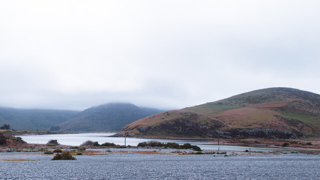 Lake Forsyth, New Zealand: Wide panoramic view of calm Wairewa lagoon water with dark shingle gravel bank and dramatic white cloud fog over hills.