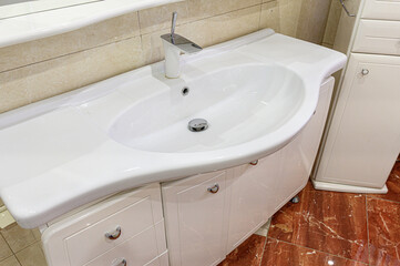 Fototapeta premium A white asymmetrical ceramic sink sits atop a glossy white vanity unit; paired with a chrome faucet, beige tiled walls, and a rich brown marble floor