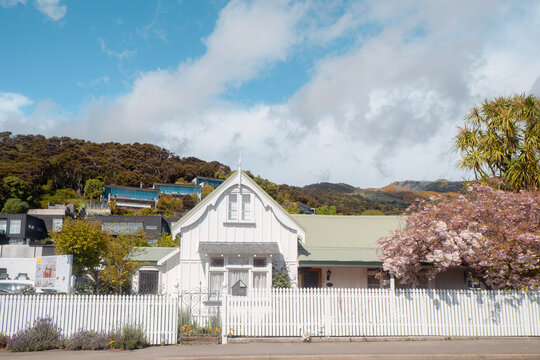 Akaroa, New Zealand: Beautiful French colonial white timber cottage with a picket fence and pink spring flowers below steep green forested hills.