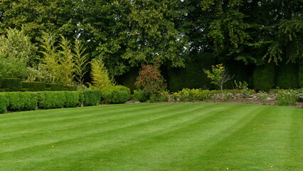 A perfectly mown green grass lawn displaying prominent mower stripes stretches across a lush...