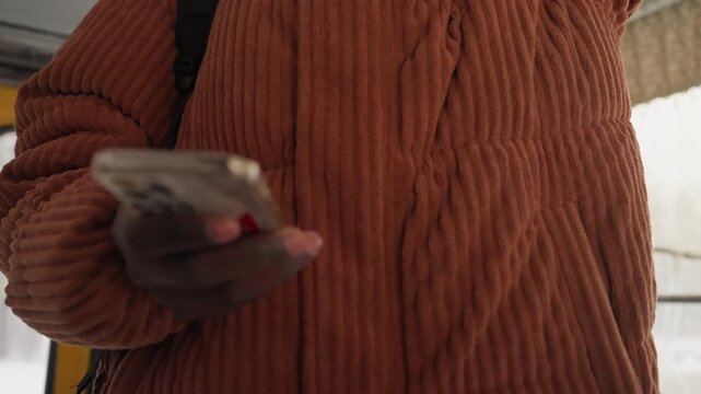 Man standing in bus holding phone and gripping rail, orange knit sweater with beanie and sunglasses, backpack strap visible, scrolling and tapping while bound for work, confident urban style, candid