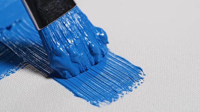 Slow motion tracking macro shot of paintbrush spreading blue acrylic paint across white textured canvas on studio tabletop with glossy wet strokes
