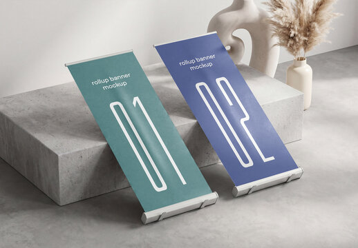 Two vertical roll-up banner mockups leaning against a concrete block podium platform in a minimalist interior setup with a ceramic vase and pampas grass background