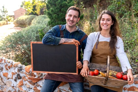 Two people in aprons holding a chalkboard and basket of vegetables outdoors.