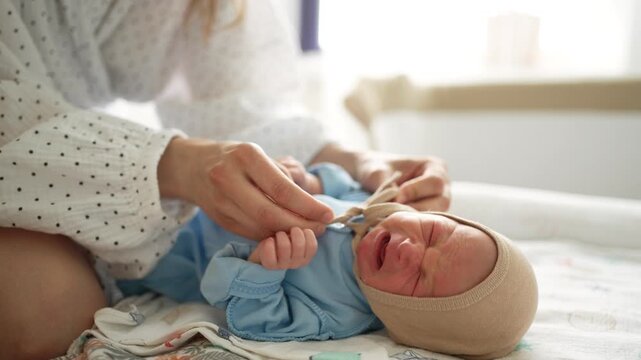 Careful mother dressing her newborn infant in a bright, sunlit room. The crying baby lies on a changing table while the woman puts on a warm blue outfit and a soft beige bonnet