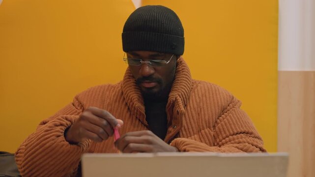 Black man opening small snack during break in yellow booth, unwraps candy near laptop and coffee cup, focused on hands with calm expression, casual coat and beanie, soft mall ambience and relaxed