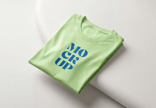 Folded light green t-shirt mockup placed on a white organic podium shape in a minimalist studio setting