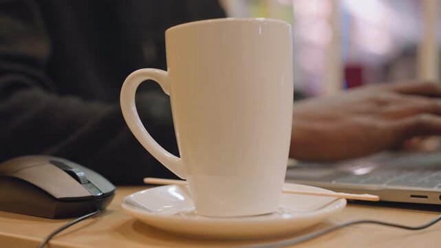 White mug on desk foreground, blurred laptop and hands typing in background, warm indoor light, saucer and mouse visible, freelance writer at laptop creating calm productive atmosphere