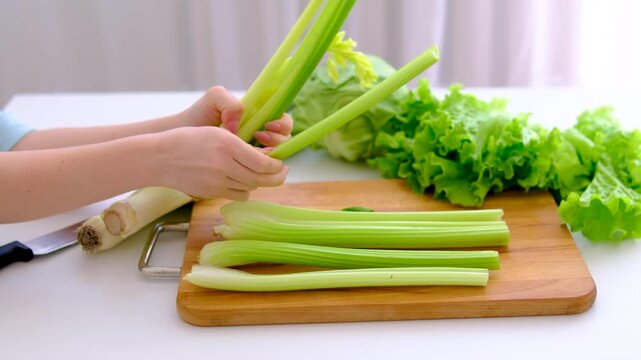 Breaking fresh celery stalks on wooden board with cabbage greens and leek in kitchen