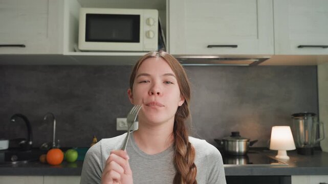 Young woman tasting food in kitchen, testing fork, braided hair, modern countertop, microwave and stove in background, oranges on counter, soft lamp glow, contemplative expression, recipe experiment