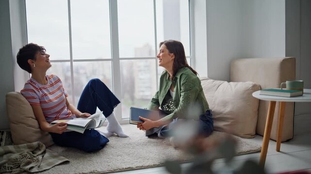 Joyful friends speaking home floor discussing literature. Brunette holding book