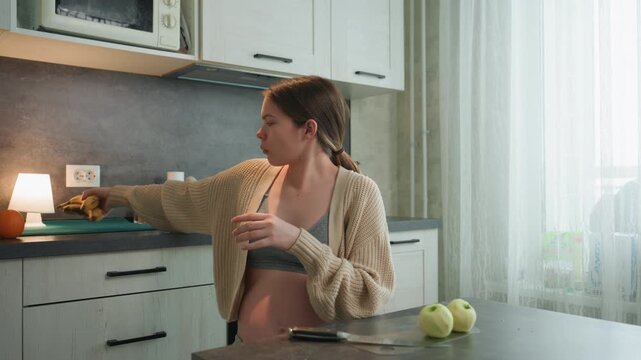 Pregnant white woman at counter reaching for banana and apples, wearing knitted cardigan and bralette, visible baby bump, warm sunlight through window, placing fruit near phone, playful expression