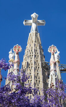 28 de mayo de 2026. Detalle de torres y cruz De la Torre central de la Sagrada Familia de Gaud&iacute; en Barcelona, y flores violetas de los &aacute;rboles del parque.