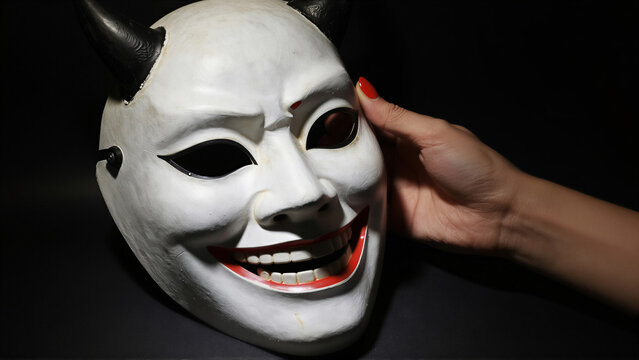 Close-up of a white demon mask with black horns held by a hand. The creepy devil face features a wide grin against a dark background for Halloween horror