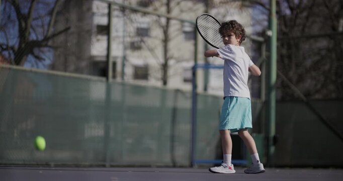 A young boy in a white shirt and blue shorts plays tennis on an outdoor court, showcasing a forehand stroke with determination.